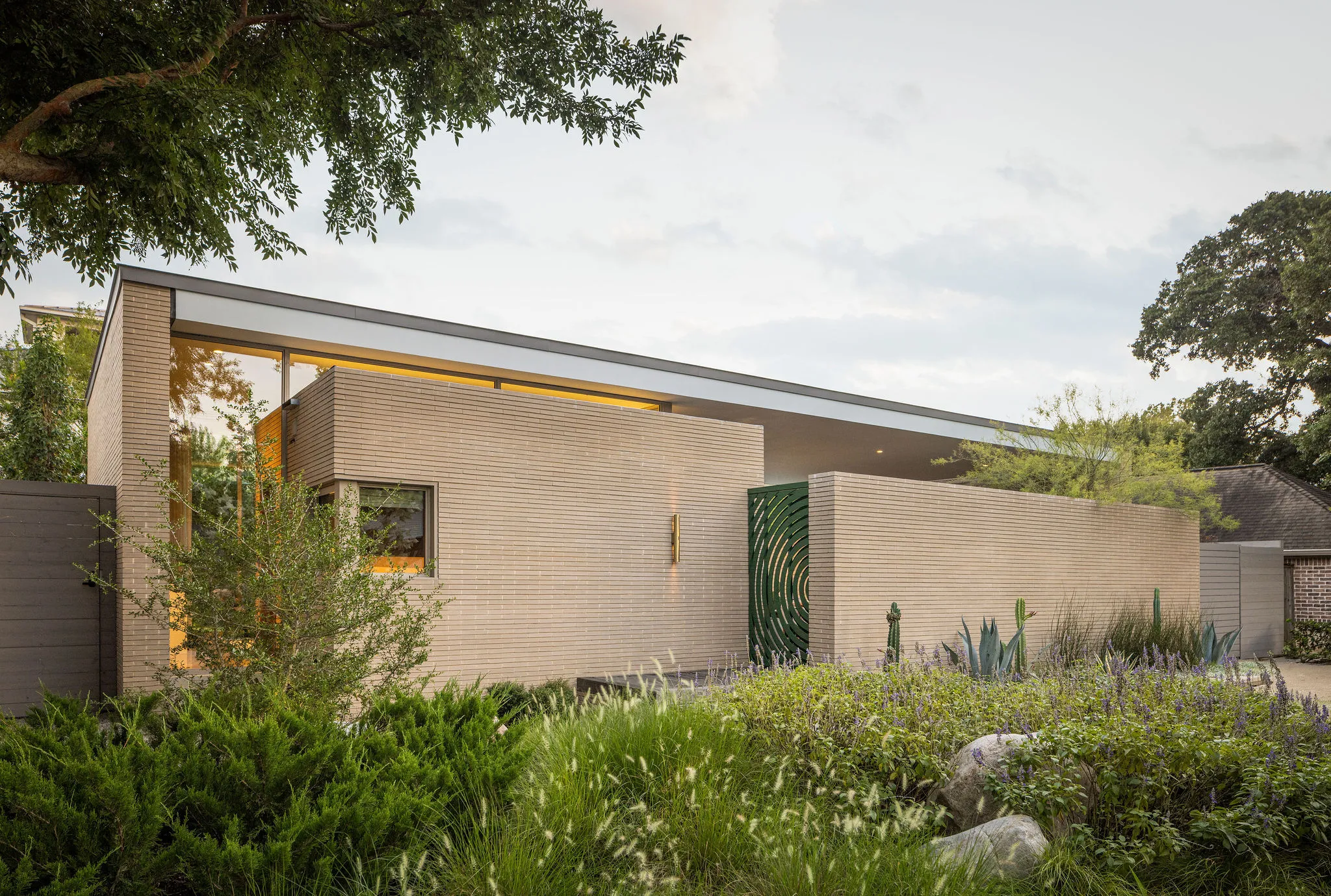 Brick street facade with a clerestory glow beneath the cantilevered roof and a green sculptural gate